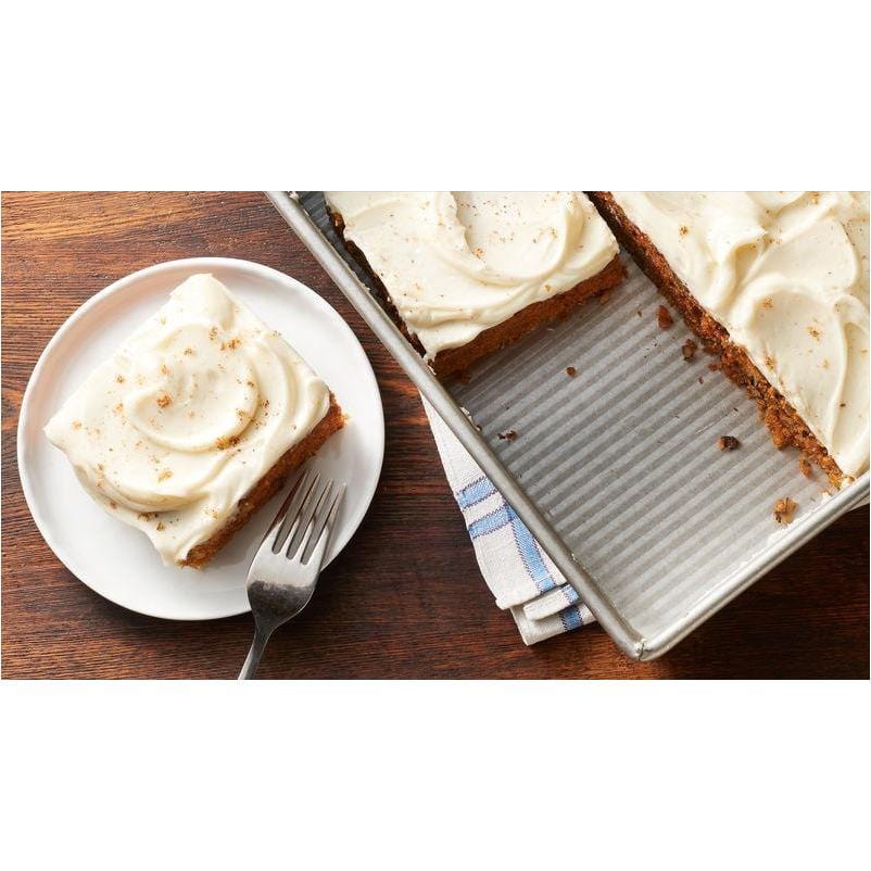 A gauche un morceau de cake au glaçage blanc sur une assiette blanche et à droite le reste du cake dans un récipient gris. Le tout sur une table en bois