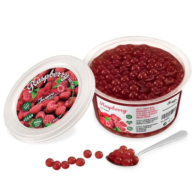 Une boite sur fond blanc remplie de perles bordeaux et à gauche le couvercle avec plusieurs framboises. Devant des perles bordeaux dans une cuillère