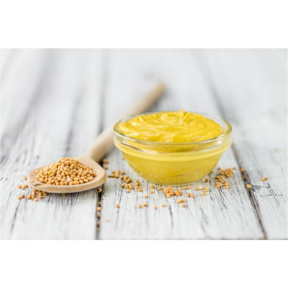 A gauche une cuillère en bois avec des graines de moutarde et à droite un petit récipient avec une sauce jaune. Le tout sur une table en bois grise