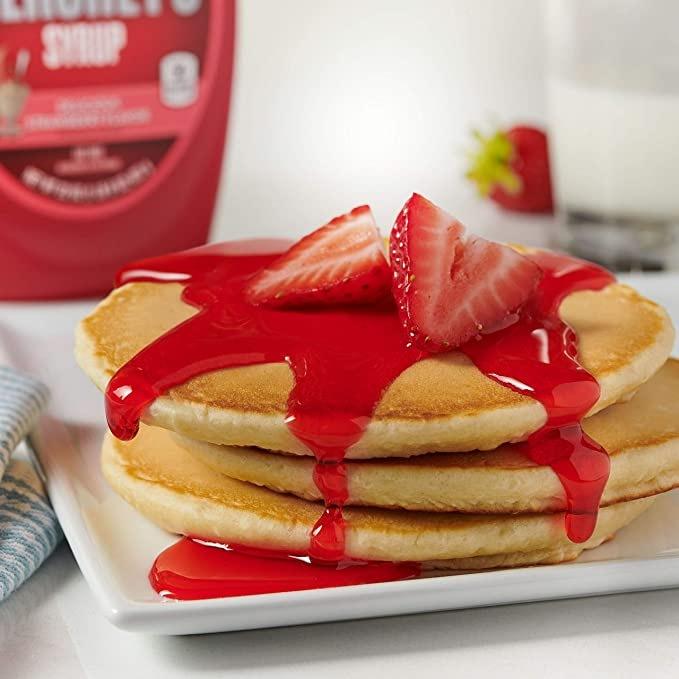 3 pancakes dans une assiette carré blanche avec au-dessus du coulis rouge et une fraise coupé en 2, à l’arrière on voit une partie de la bouteille rouge