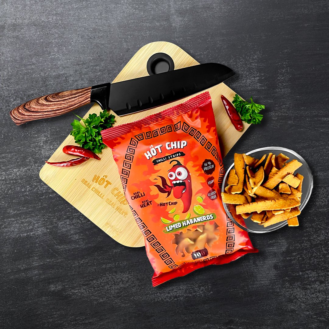 Un paquet rouge sur une planche en bois avec un couteau noir et à droite un bol avec des chips de forme rectangulaire. Le tout sur une table noire