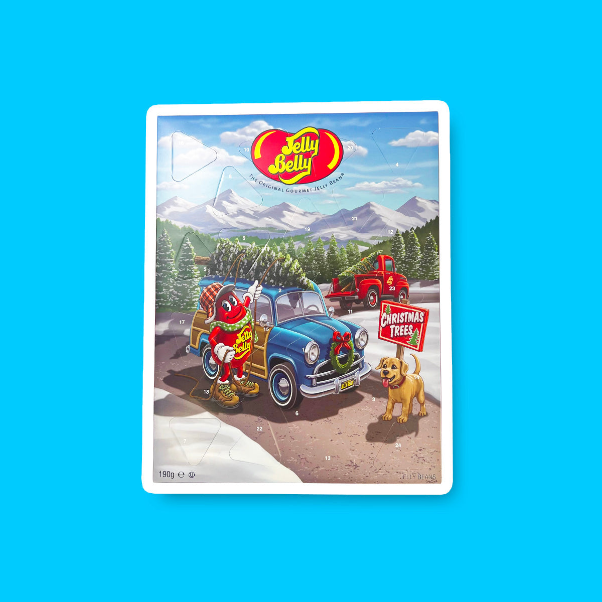 Un carton sur fond bleu avec une voiture bleue autour d’un paysage montagneux et enneigé avec plein de sapin. Sur le devant, un chien et un Jelly Belly rouge qui attache un sapin sur le toit de sa voiture