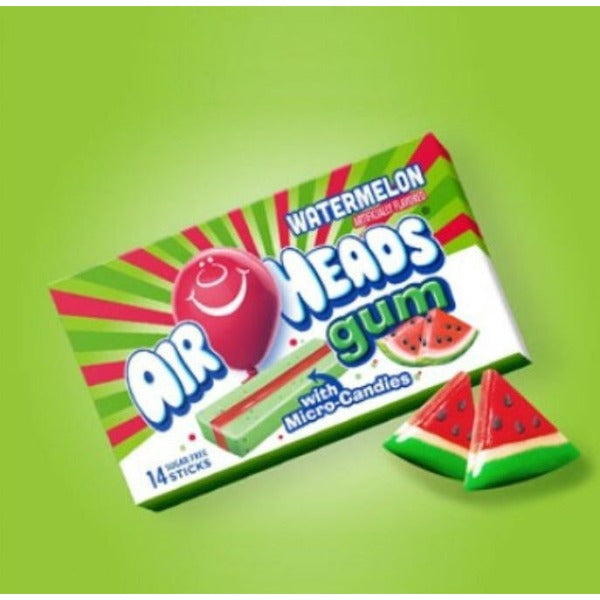 Un paquet rouge-vert sur fond vert avec un ballon rouge qui sourit et il y a un chewing-gum rouge/vert et à droite 2 morceaux de pastèque. Devant il y a 2 jouets en forme de pastèque 