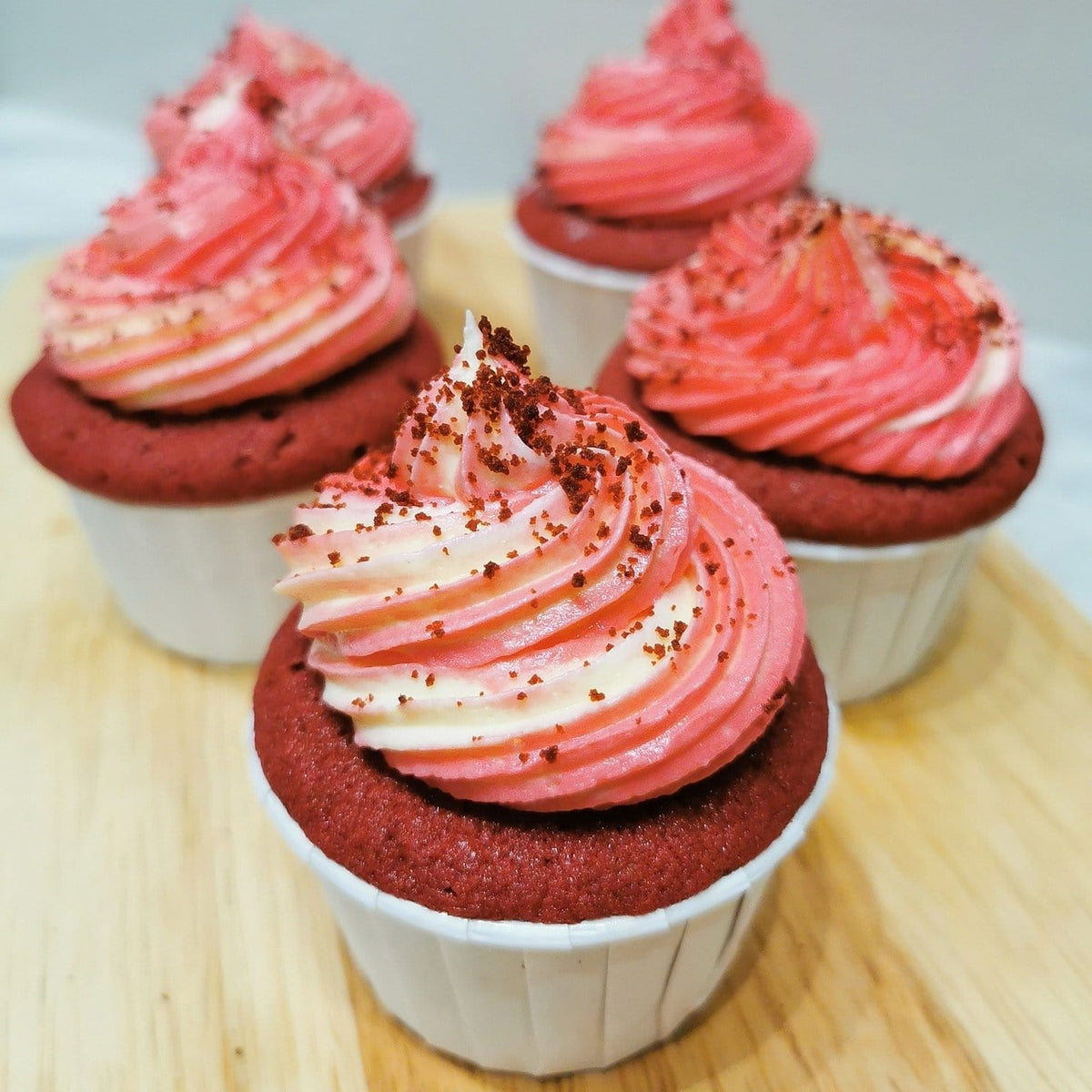 5 petits cupcakes rouges dans de petits papiers blancs avec un glaçage blanc et rose, le tout sur une planche en bois
