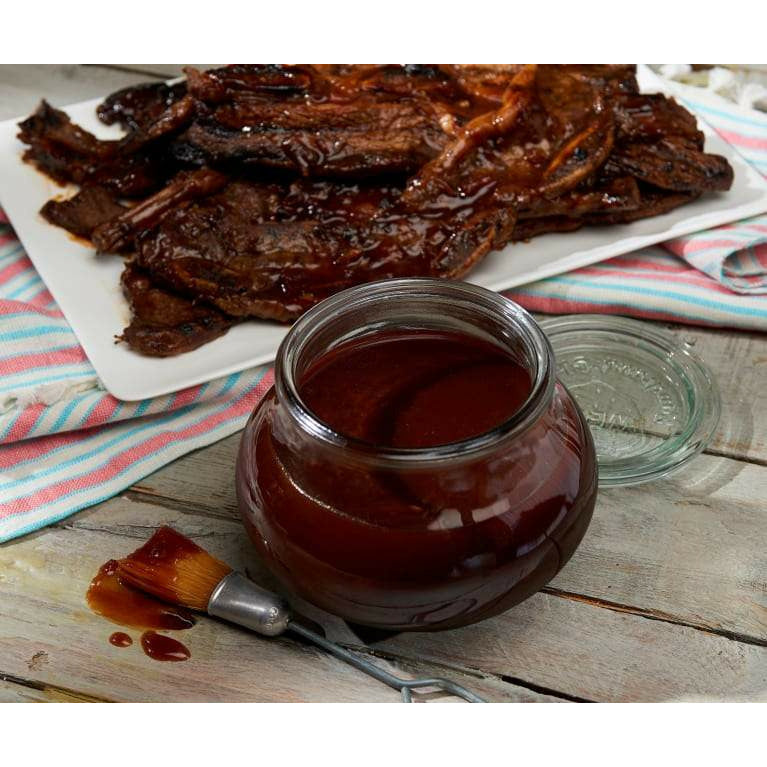 Un bocal rond en verre avec une sauce rouge, à l’arrière de la viande dans une assiette blanche et à coté un pinceau avec de la sauce. Le tout sur une table en bois 