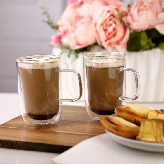 2 tasses transparentes de boissons brunes sur une planche en bois. Devant une assiette blanche avec des pâtisseries et derrière des fleurs roses