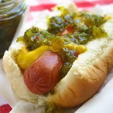 Un hot-dog avec une saucisse rouge et au-dessus une sauce jaune et une sauce verte avec des morceaux vert et rouge 