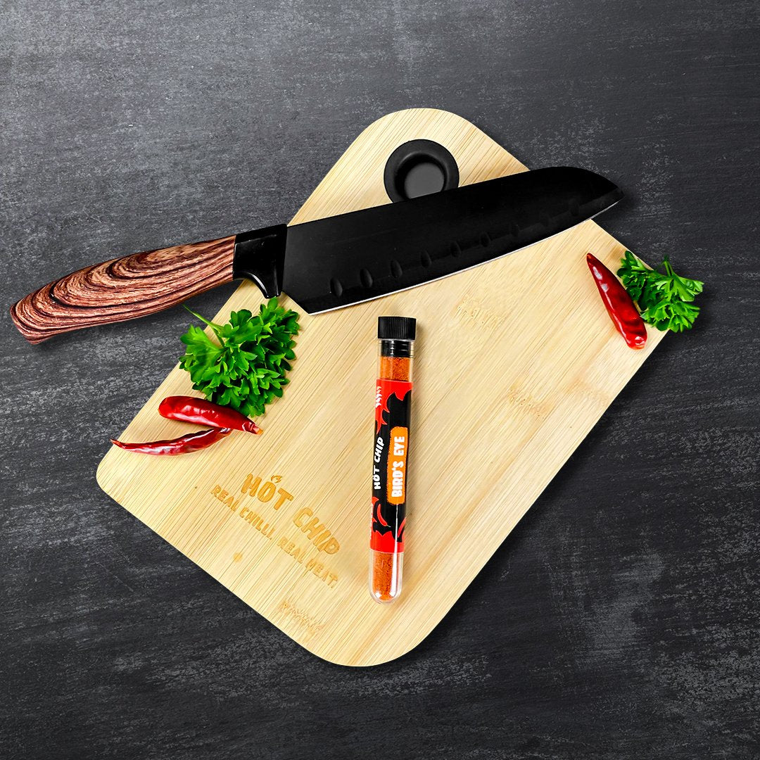 Une planche en bois sur une table noire avec au-dessus un couteau noir et une fiole en verre avec des épices rouges