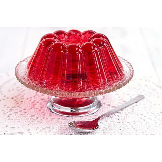 Une gelée rose sur un plat à dessert en verre et devant une cuillère en argent, le tout sur une table blanche avec une nappe en dentelle blanche