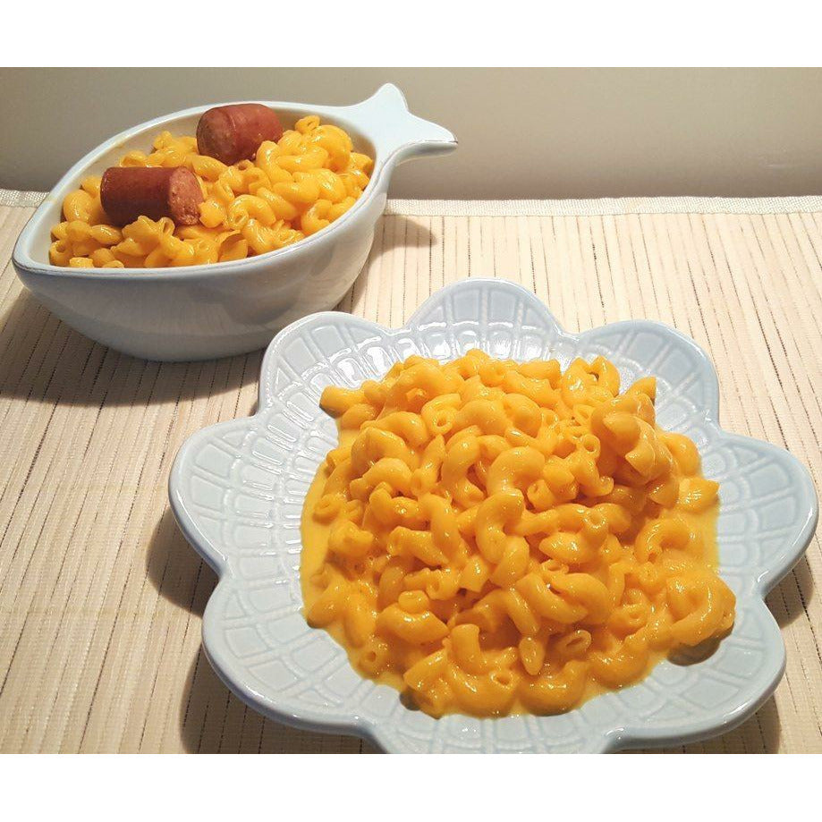 Une assiette blanche rempli de macaroni jaune et derrière à gauche un plat en forme de poisson rempli de macaroni et 2 morceaux de saucisses. Le tout sur une table en bois