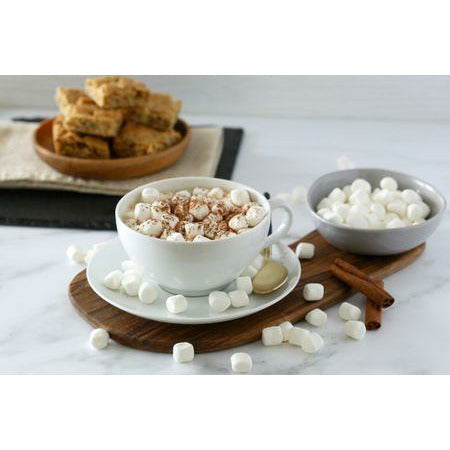 Une tasse blanche avec du chocolat chaud et plein de petit marshmallows dans un bol gris. Derrière une assiette en bois avec 6 morceaux de cakes