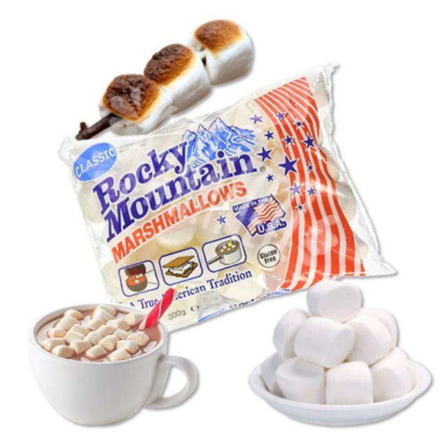 Un grand paquet transparent de marshmallows blancs au centre, en bas à gauche une tasse de cacao avec les marshmallows, en bas à droite une assiette blanche de ces marshmallows et au-dessus des marshmallows passés au feu