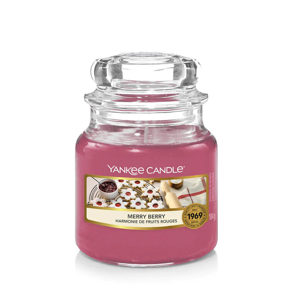 Un emballage sur fond blanc avec une bougie mauve dans un bocal en verre, un capuchon en verre et une étiquette avec des biscuits en forme d’étoiles et un pot rempli d’une sauce rouge